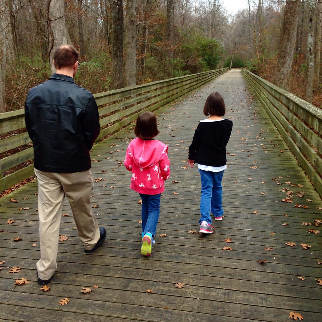 Image is of a man and two young girls taking a family walk down a long wooden bridge leading onto a gravel walking trail in a wooded area.